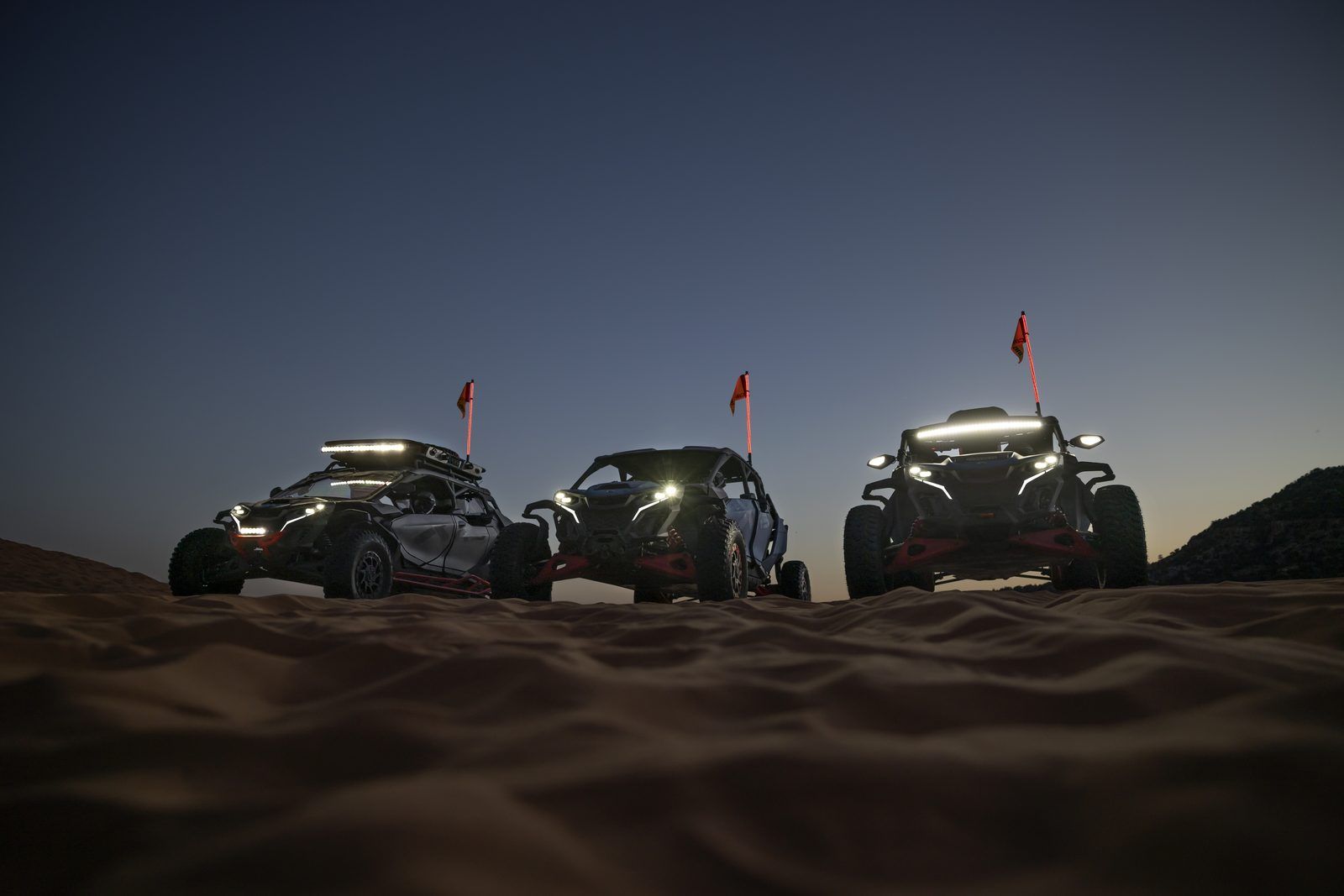 Three four-seat Can-Am Maverick X3 UTVs parked on dunes at dusk with lights on
